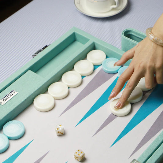 Hand placing white piece on teal and white BacoBackgammon board with dice and blue pieces, cup in background