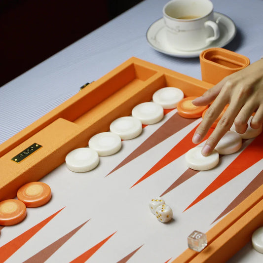 Luxury Baco backgammon set with orange and white vegan leather, white and orange pieces, hand moving a piece, dice, and a white teacup on table